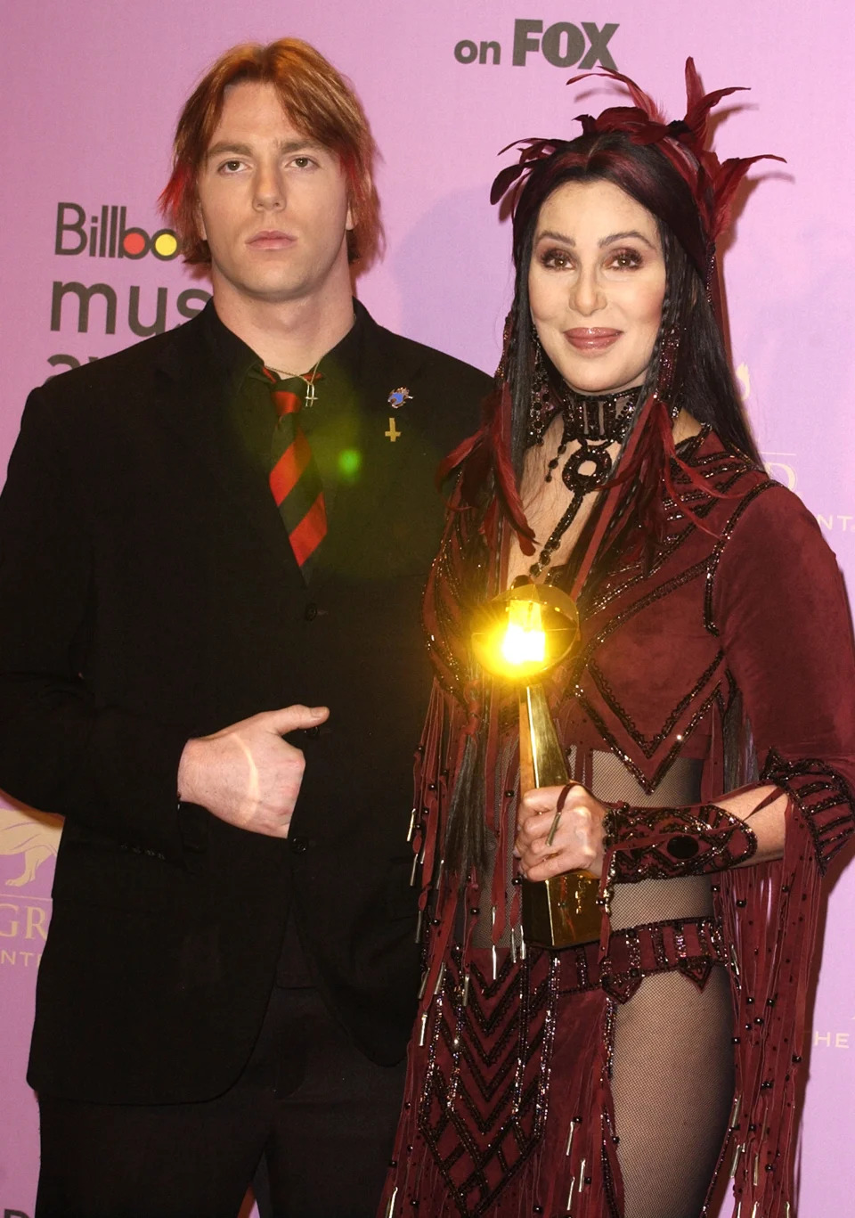 Cher in an elaborate outfit holds a Billboard Music Award next to an individual in a black suit with a red and black striped tie at a Billboard Music Awards event