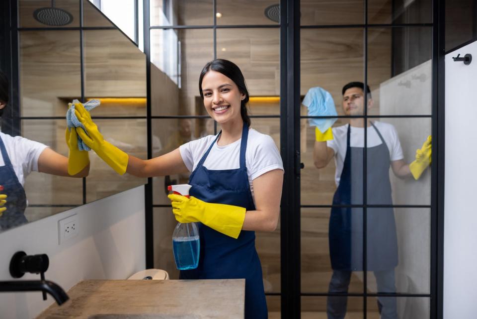 A smiling woman in yellow gloves wipes down a bathroom mirror while a man cleans a shower behind her.