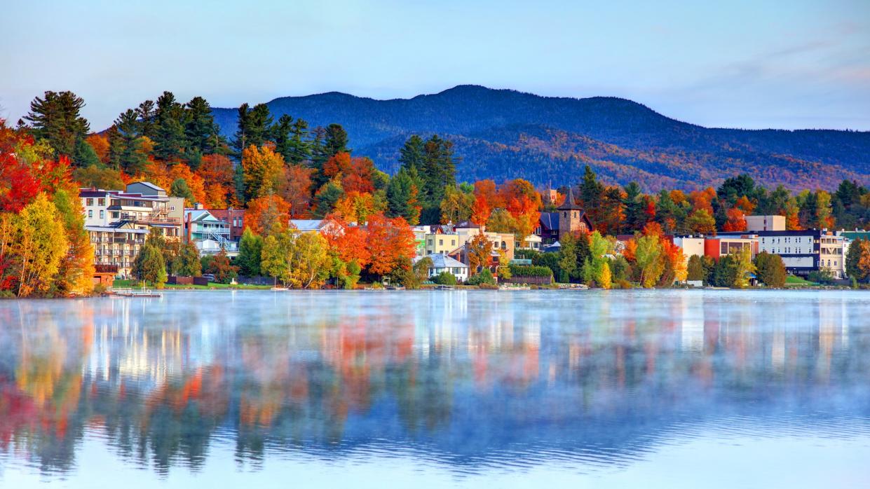  A large body of water surrounded by autumnal trees with vibrant orange and yellow leaves with houses nestled between them. 