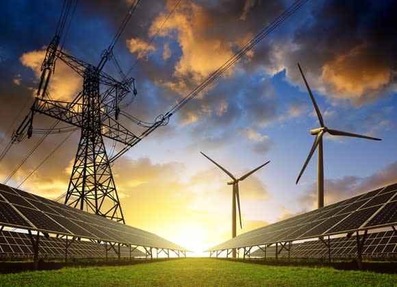 Solar panels with wind turbines and electricity pylon at sunset.