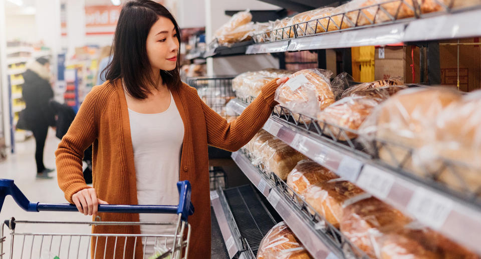 A woman chooses bread in supermarket aisle.
