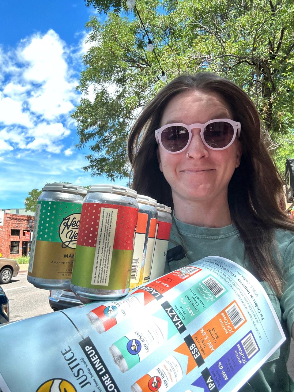 A woman wearing sunglasses holds several packs of craft beer cans outdoors, smiling at the camera