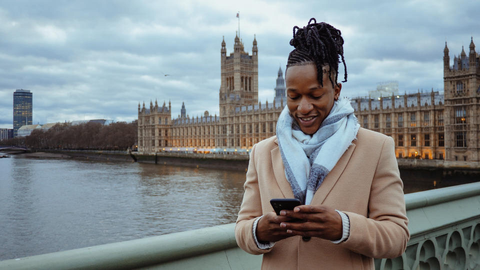 A person stands on a bridge overlooking the River Thames and the Houses of Parliament, smiling at their phone, wearing a coat and scarf