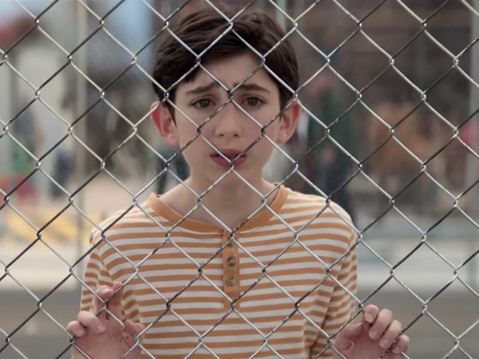 a child standing on the other side of a chain link fence with a worried expression on his face