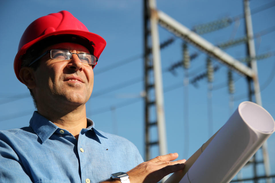 A man in a hard hat with blueprints and high voltage power lines behind him
