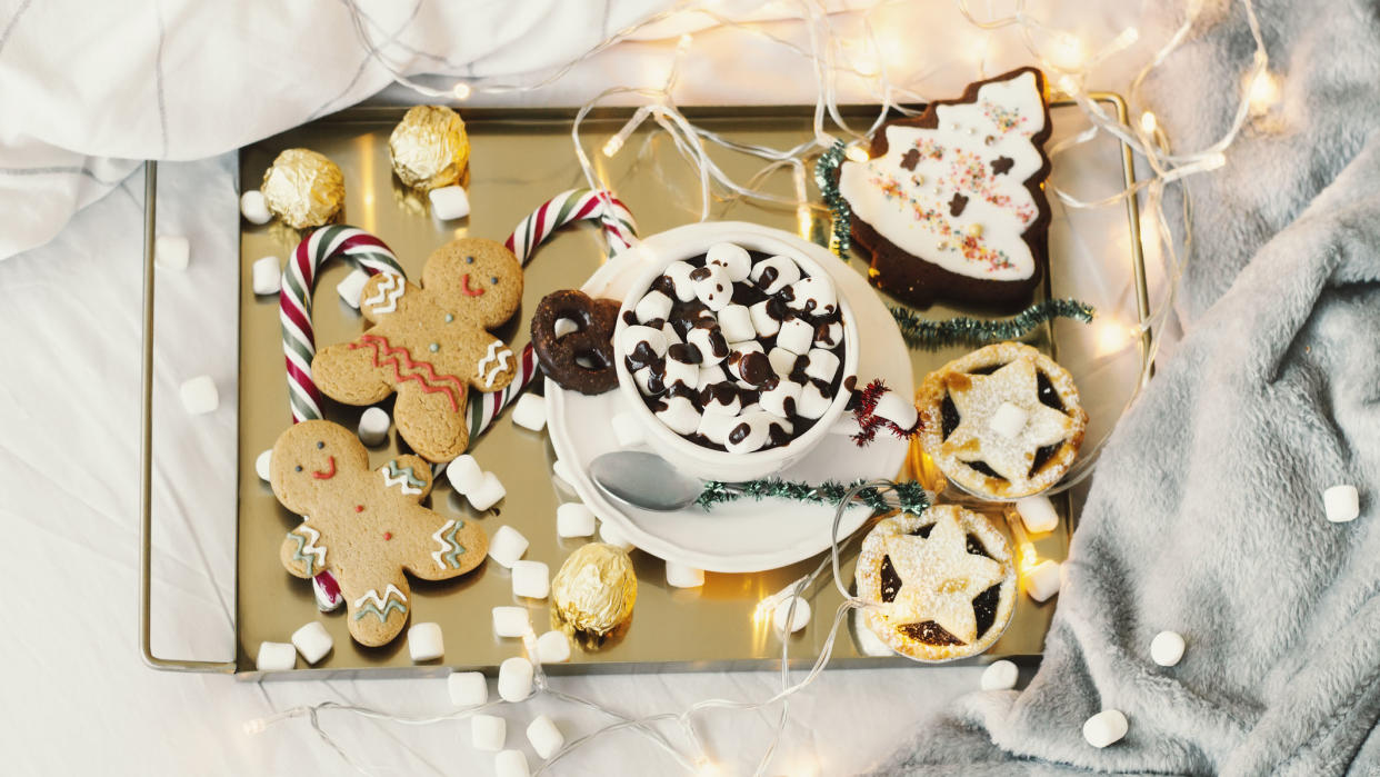  The the best (and worst) festive foods for sleep image shows a golden tray of gingerbread cookies, mince pies and marshmallow-topped hot chocolates placed on a bed dressed with faux fur blankets. 