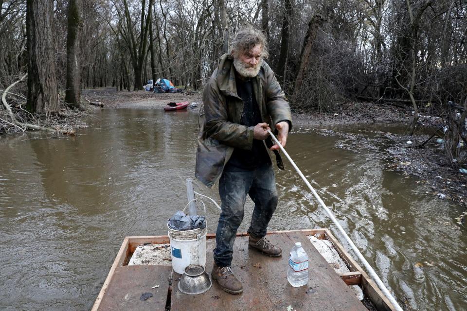 bearded man wearing rain jacket pulls rope on wooden raft in flooded forest river with tent in background