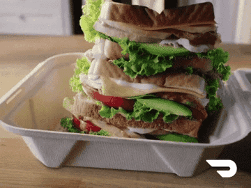 A large stacked sandwich with multiple layers of bread, lettuce, tomatoes, and other fillings, placed in a takeout container with the DoorDash logo in the bottom right corner