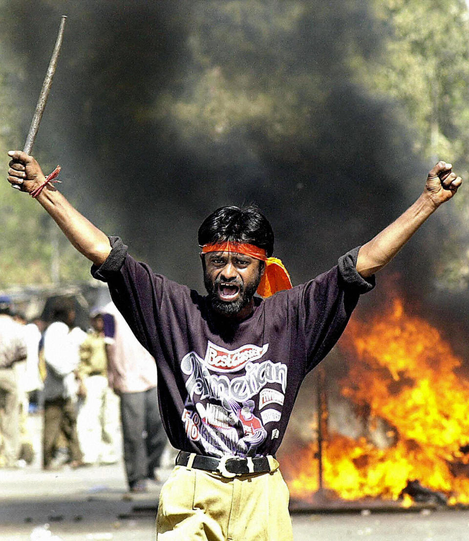 A man wearing a red scarf tied around his head and holding an iron rod raises his hands triumphantly against a backdrop of flames and smoke.