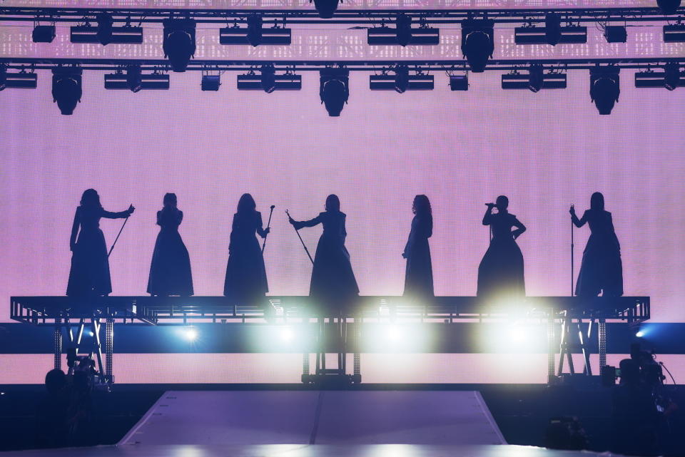 Seven women stand on an elevated stage in silhouette, each holding a microphone. The background is a large, brightly lit screen