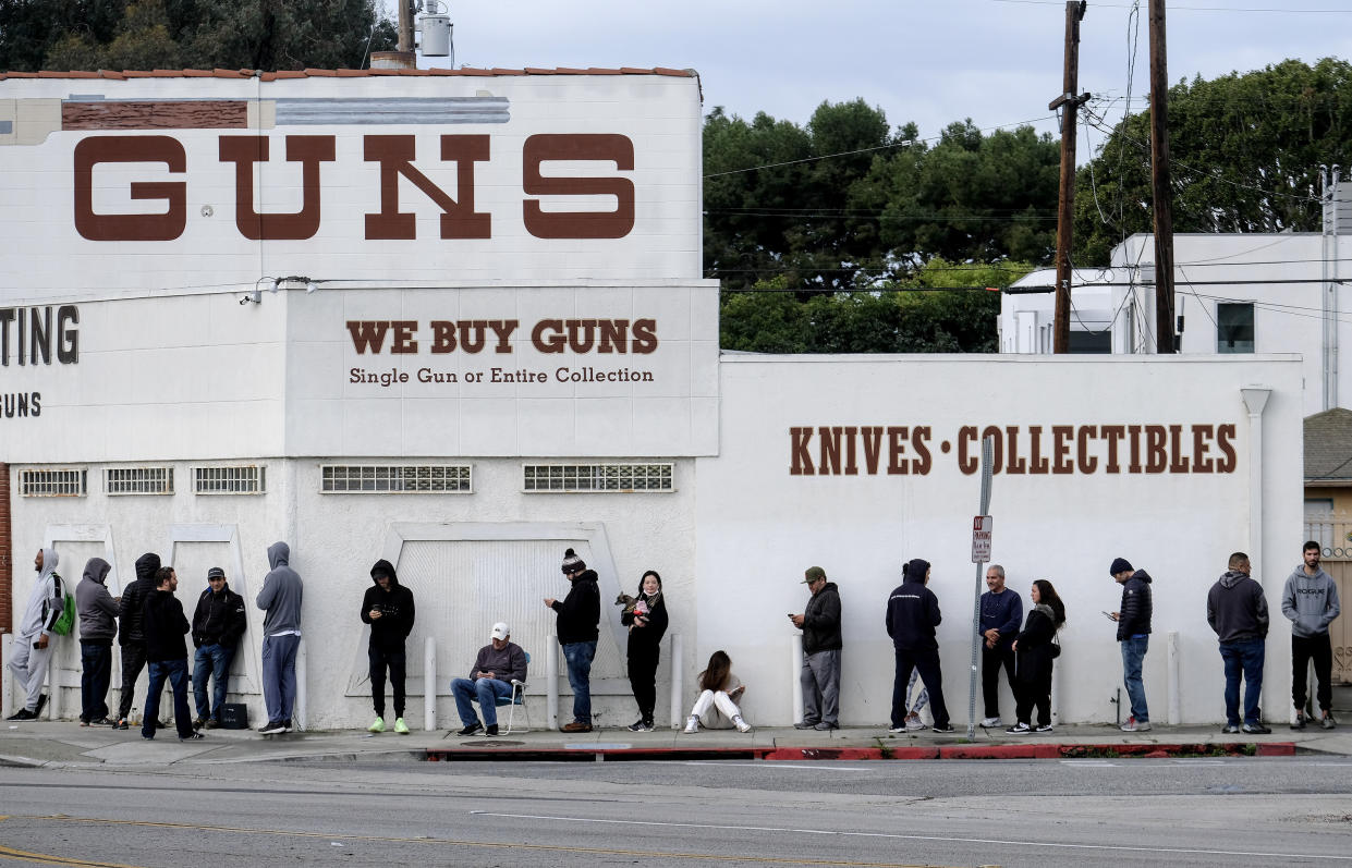 A long line of people standing and seated on folding chairs and on the ground, wait to enter a store marked: GUNS, Knives, Collectibles, and We Buy. Guns, Single Gun or Entire Collection.