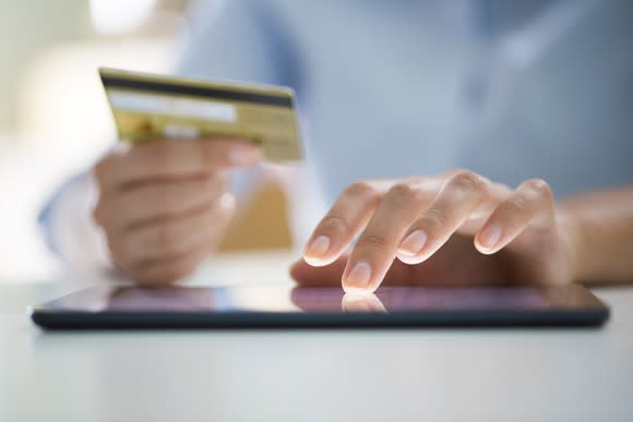 A woman holding a credit card with her right hand as she's shopping online using a tablet.
