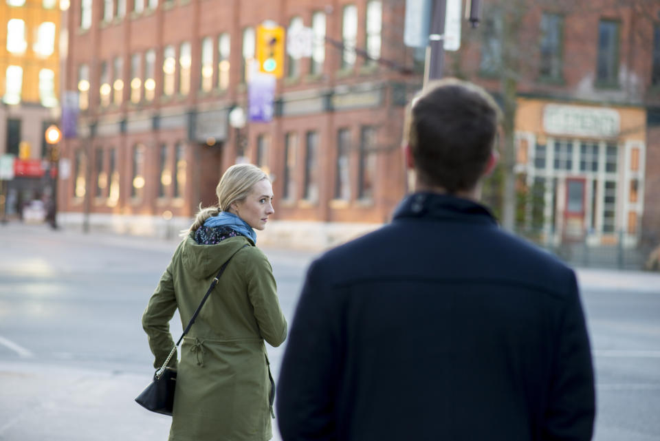 Scared young woman with blonde hair looking back over her shoulder at a stranger in a black trench coat that is following behind her downtown in an urban city.