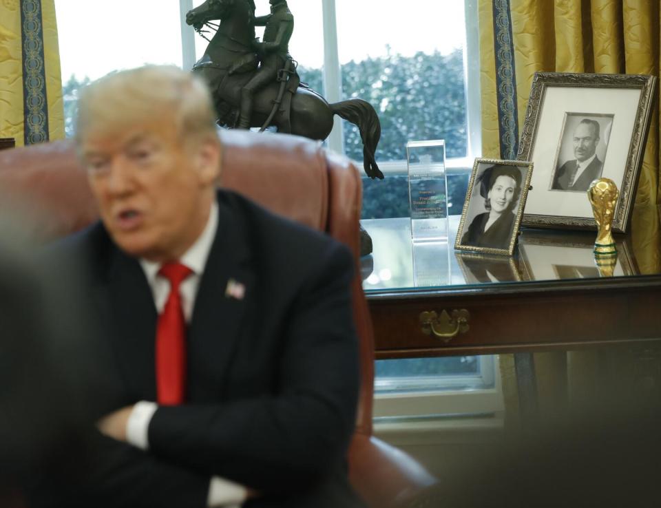 A man in dark suit and red tie, arms folded, speaks in front of a desk with framed photos and a statue of a person on a horse