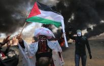 <p>A Palestinian man holding a flag walks in the smoke billowing from burning tires next to a protester wearing an Anonymous mask during clashes with Israeli forces along the border with the Gaza Strip east of Khan Younis on May 14, 2018, as Palestinians protest the inauguration of the U.S. Embassy following its controversial move to Jerusalem. (Photo: Said Khatib/AFP/Getty Images) </p>