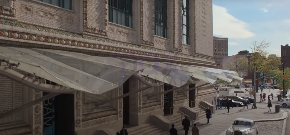 A busy sidewalk outside a large brick building with glass awnings, people walking, and cars parked along the street