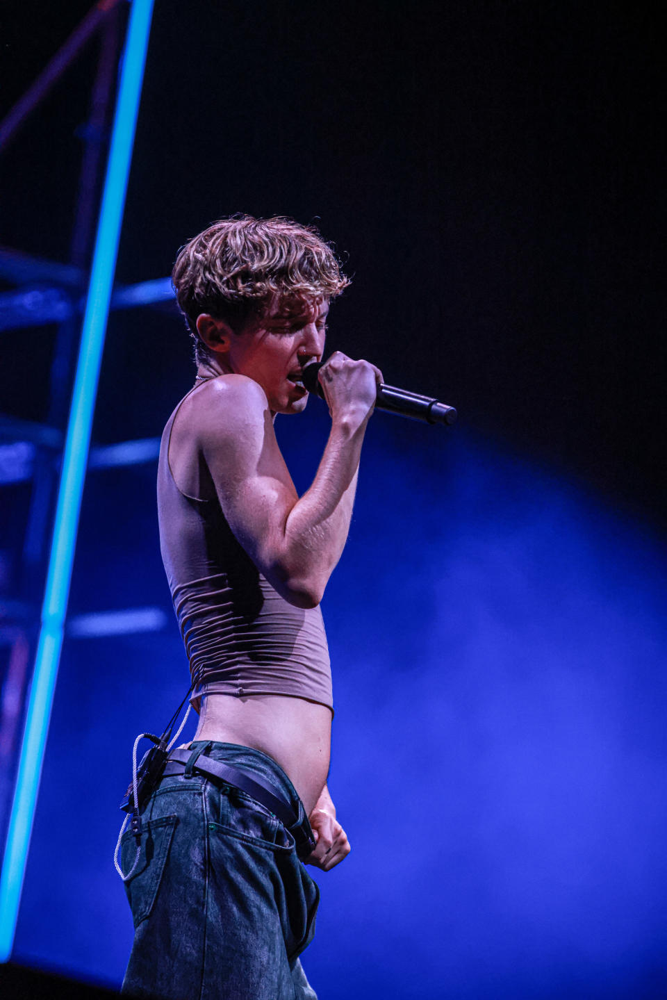 Troye Sivan performs passionately on stage, wearing a sleeveless crop top and jeans, holding a microphone close to his mouth