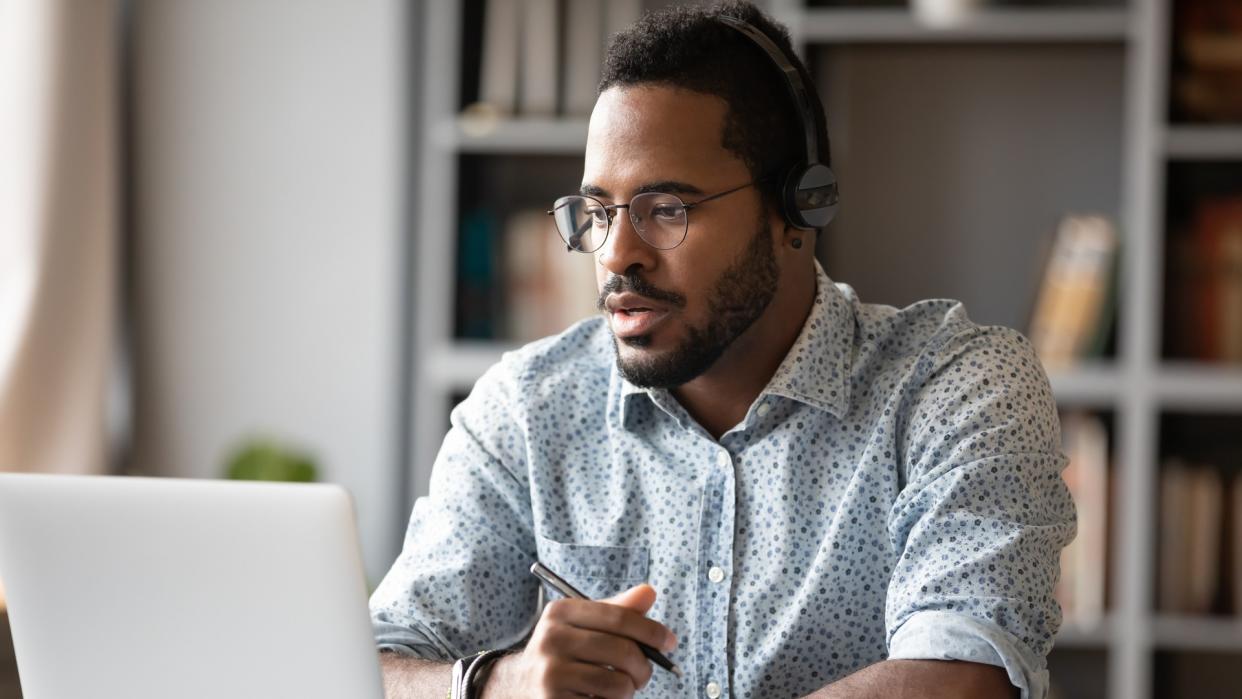 Focused young african businessman wear headphones study online watching webinar podcast on laptop listening learning education course conference calling make notes sit at work desk, elearning concept.