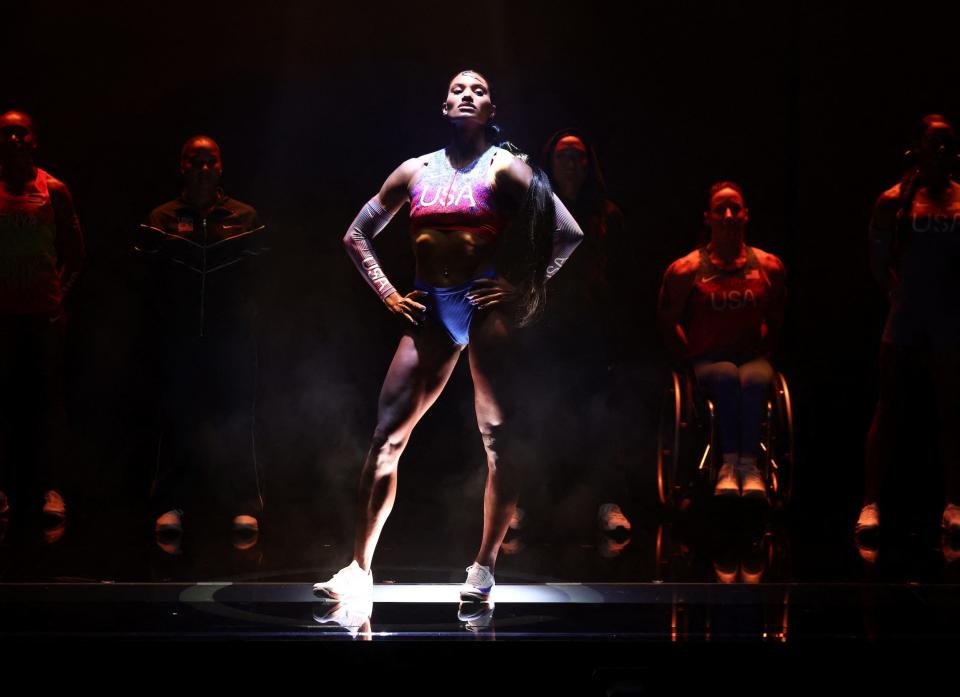 A woman poses on a runway under a spotlight wearing a red white and blue bra and high-cut blue bikini bottoms