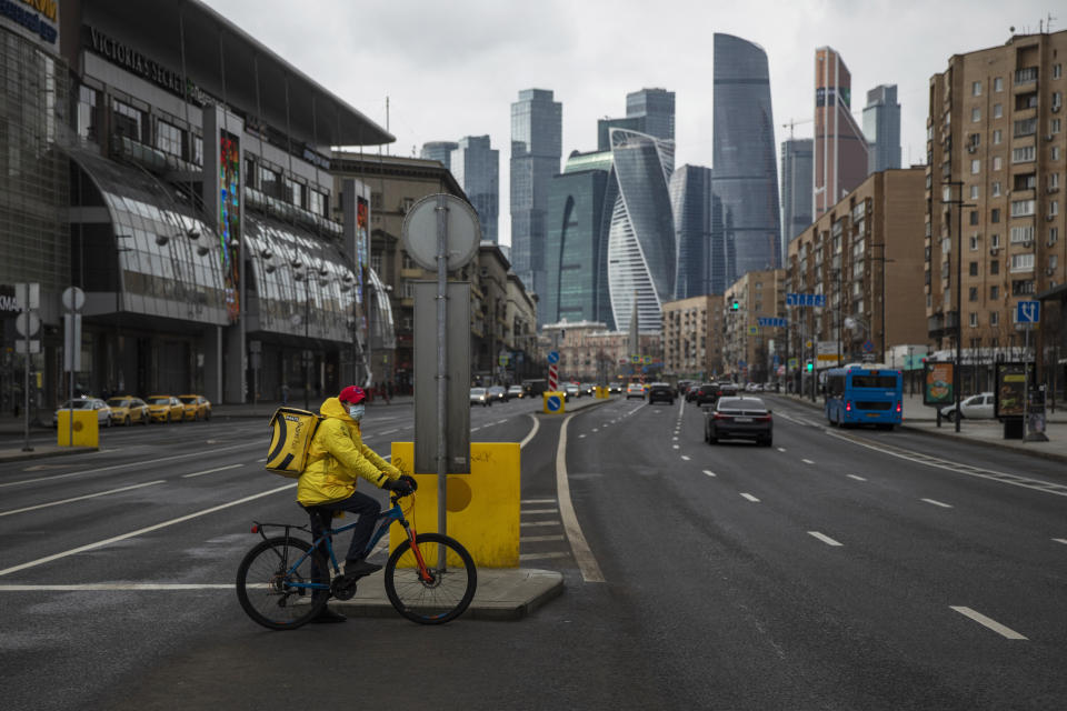 A food delivery courier wearing a face mask waits to cross a nearly empty road due to residents taking the advice of staying at home to avoid the spread of the coronavirus, with Moscow City skyscrapers in the background, in Moscow, Russia, Thursday, April 2, 2020. Online shopping for food and other supplies has boomed in Moscow after the authorities put most residents on lockdown. The new coronavirus causes mild or moderate symptoms for most people, but for some, especially older adults and people with existing health problems, it can cause more severe illness or death. (AP Photo/Pavel Golovkin)