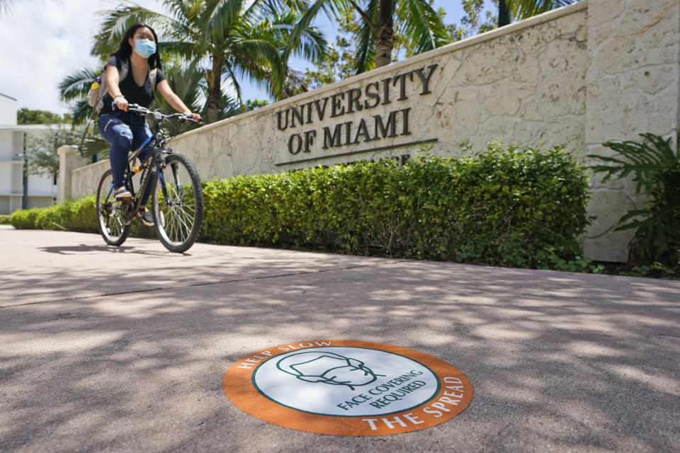 FILE - In this Aug. 25, 2020, file photo, a cyclist, wearing a mask to prevent the spread of the new coronavirus, rides by an entrance to the University of Miami in Coral Gables, Fla. The torrid coronavirus summer across the Sun Belt is easing after two disastrous months that brought more than 35,000 deaths. (AP Photo/Wilfredo Lee, File)