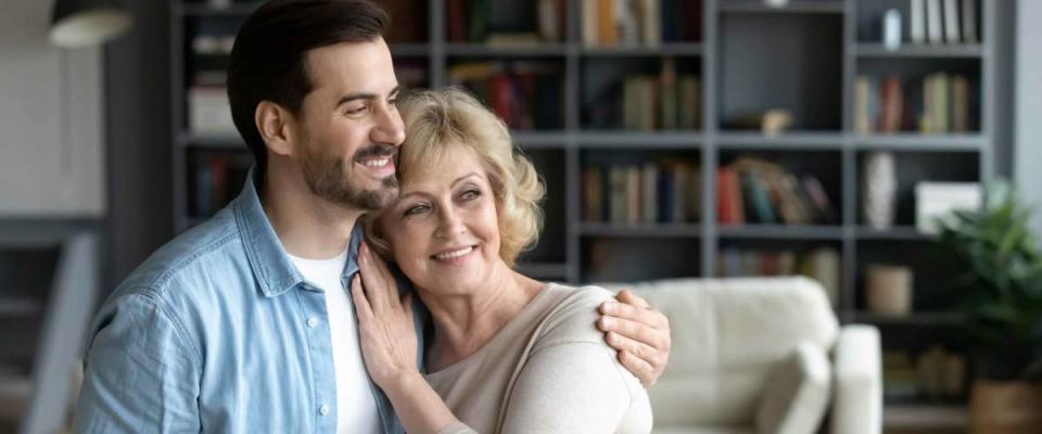 Horizontal wide image inside of cozy living room at home standing hugging middle-aged mother and grown up adult