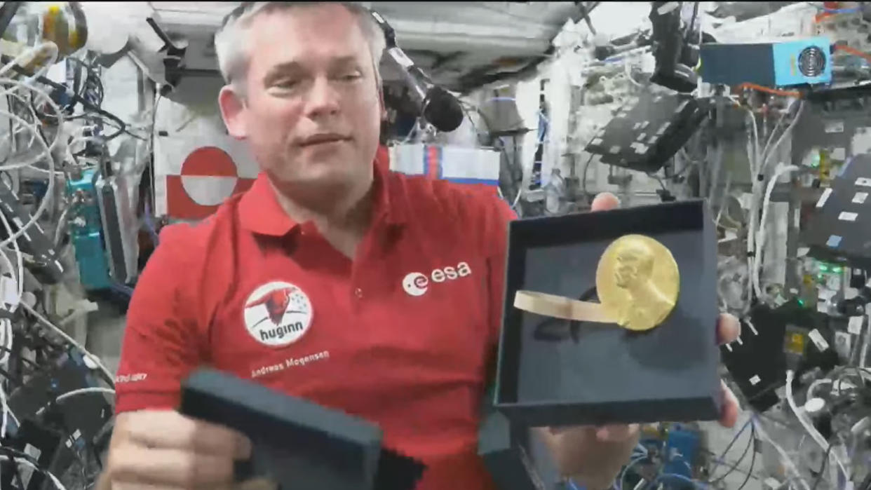  An astronaut in a red shirt holds a gold Nobel Prize medal in space. 