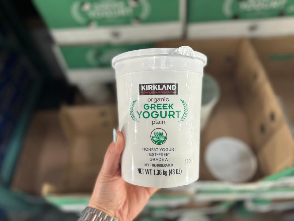 A hand holds a white tub of Kirkland Signature Greek yogurt with green lettering on it in front of cardboard boxes at Costco
