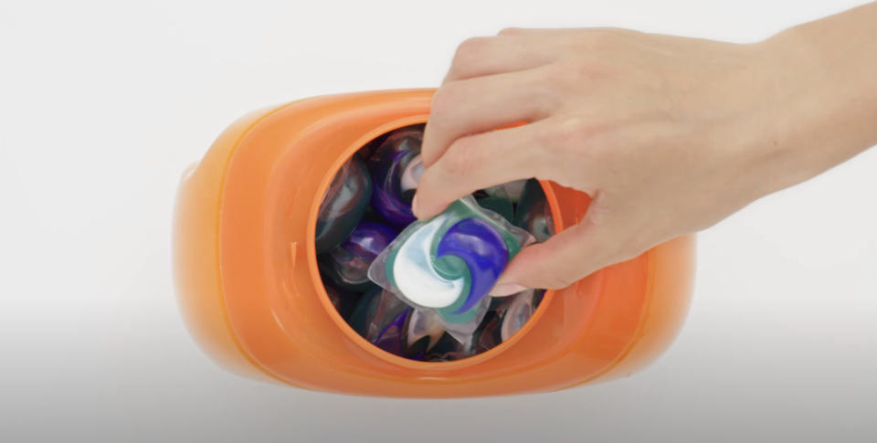 A hand placing a laundry pod into an orange container with a slot
