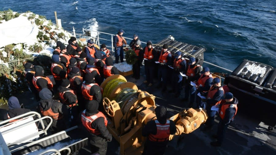 The crew of the USCG Cutter Mackinaw huddles together aboard the ship, wearing their blue uniforms and orange safety vests. A large pile of bundled Christmas trees sits at the back of the ship.