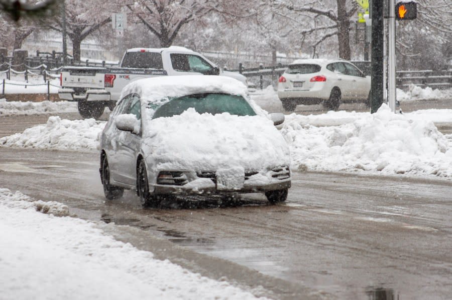 A car covered in snow with a clean windshield drives in downtown Denver.