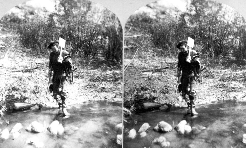 A Stereograph of a miner carrying equipment while walking across a shallow creek in Colorado in 1874.