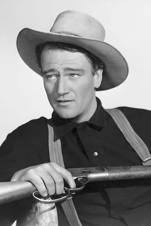 John Wayne is wearing a cowboy hat and suspenders, holding a rifle while looking into the distance