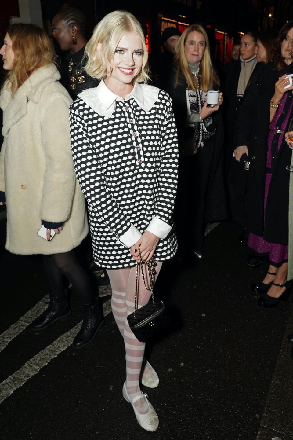 Lucy Boynton, Chanel, flats, Mary Janes, Mary Jane flats, white flats, white Mary Janes, grosgrain flats, ribbon flats, tights, sheer tights, dress, black dress, printed dress, Metiers D'Art, runway, fashion, runway show, fashion show, Manchester, England