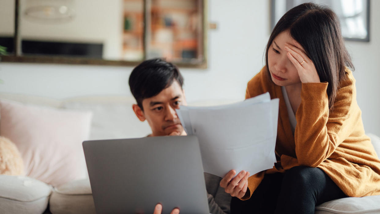  image depicts a stressed asian couple looking at pieces of paper and a laptop  