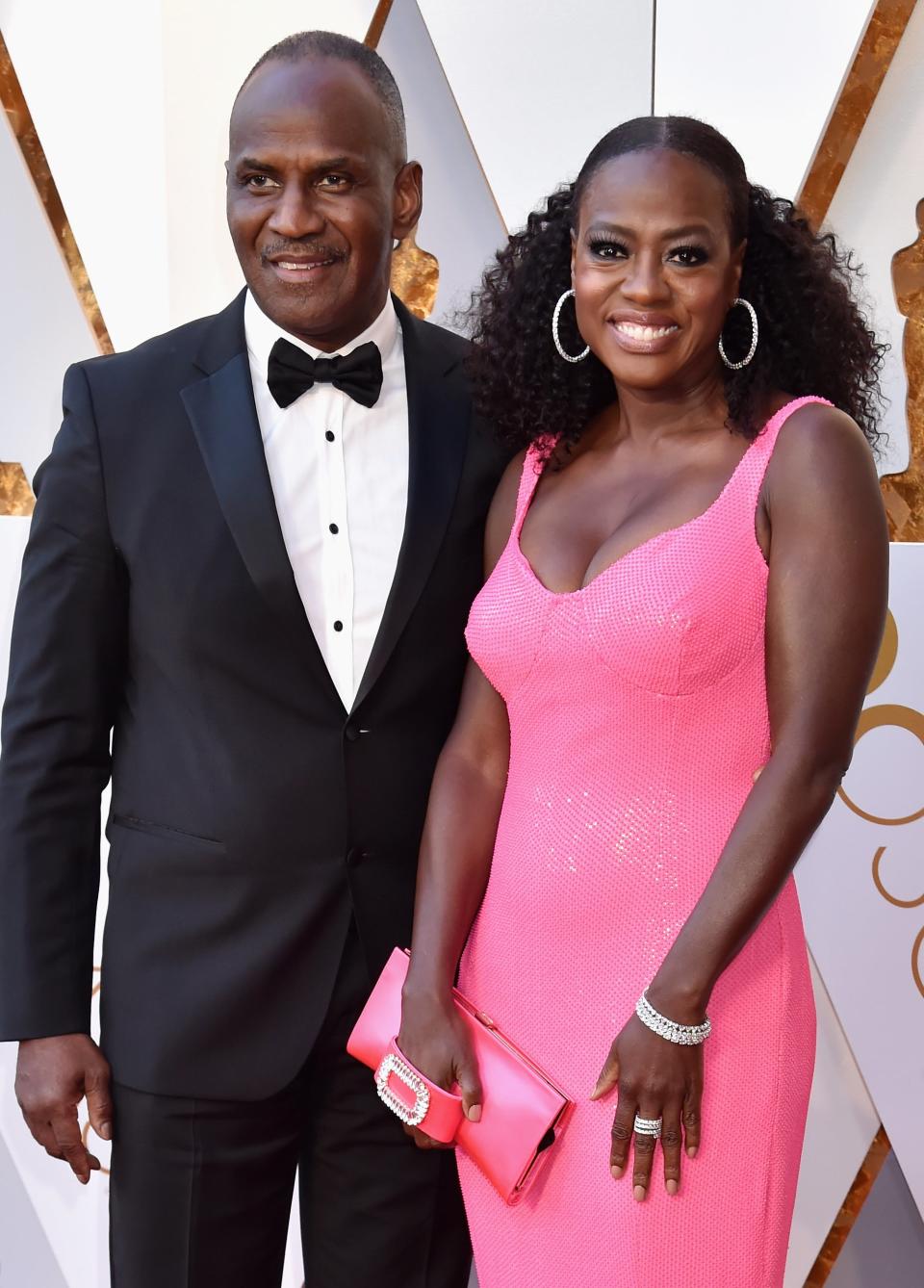 Two individuals posing at the Oscars, one in a black tuxedo, the other in a pink sleeveless gown with a clutch