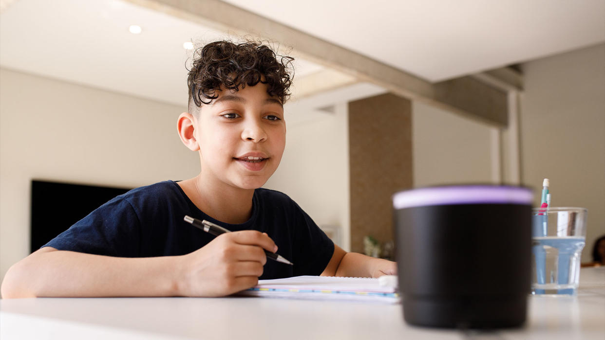  Child with curly brown hair sits at a table with a pencil and looks at a smart speaker on the table, as if to ask it a question. 