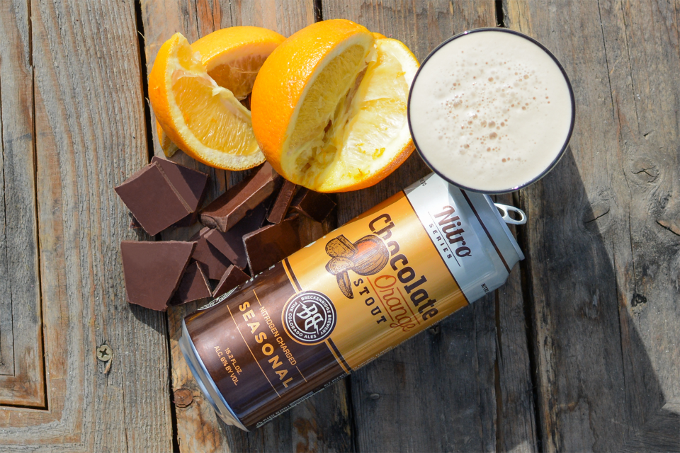 A can of chocolate orange stout beer with orange slices and pieces of chocolate, all set on a wooden surface.