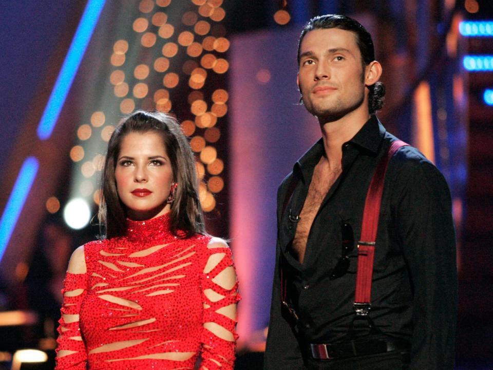 Kelly standing in a red strappy dress and Alec in a black shirt and red suspenders.
