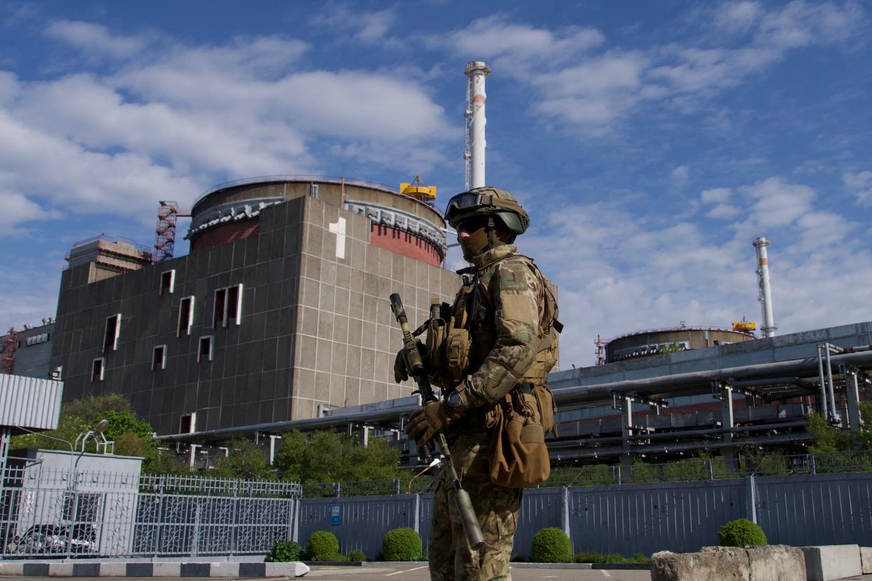 A Russian serviceman wearing fatigues and a helmet and holding a rifle stands near industrial buildings.