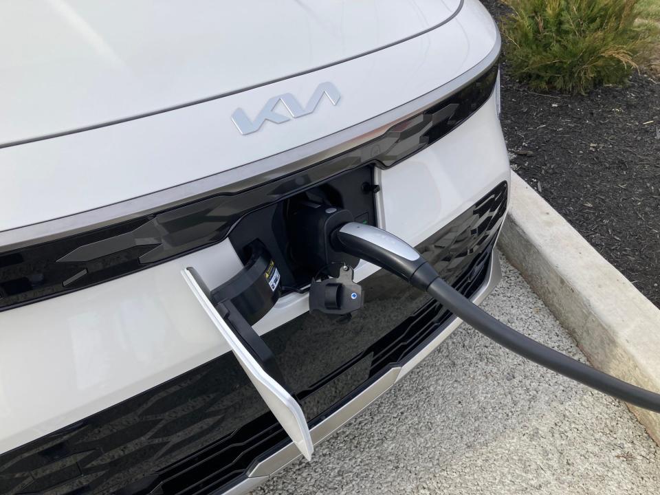 The front end of a white Kia Niro EV electric SUV with a Tesla charging cord inserted into its charge port.