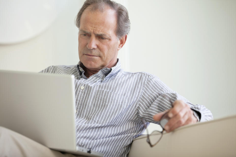 A senior man sitting on a couch and closely reading material on his laptop.