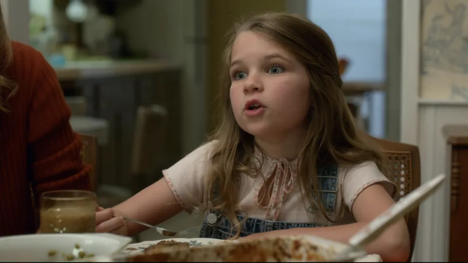 Young Missy at a dinner table in a scene looking surprised, wearing a denim dress with a blouse