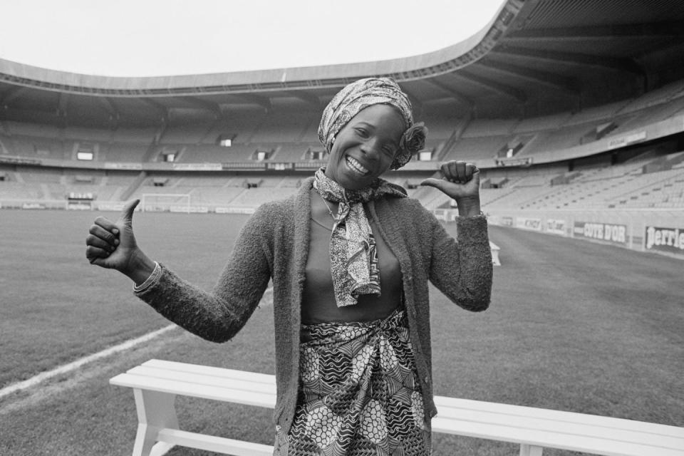 rita marley smiles at the camera and gives two thumbs up with her arms extending by her sides, she stands inside an empty stadium wearing a patterned head scarf, neck scarf and skirt with a long sleeve sweater over her shirt