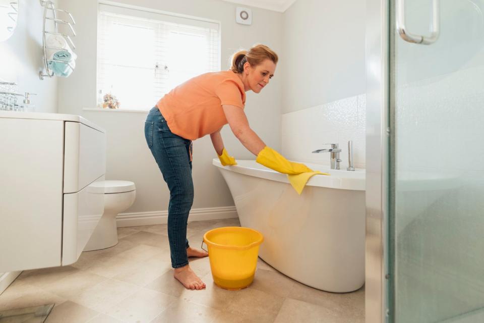 A mid-adult caucasian woman wearing protective yellow gloves is cleaning the bathtub with soap and water in her bathroom at her home in Hartlepool.