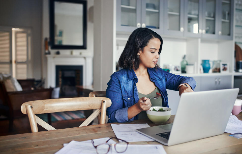 Shot of a young woman checking her watch and having a salad while working from home