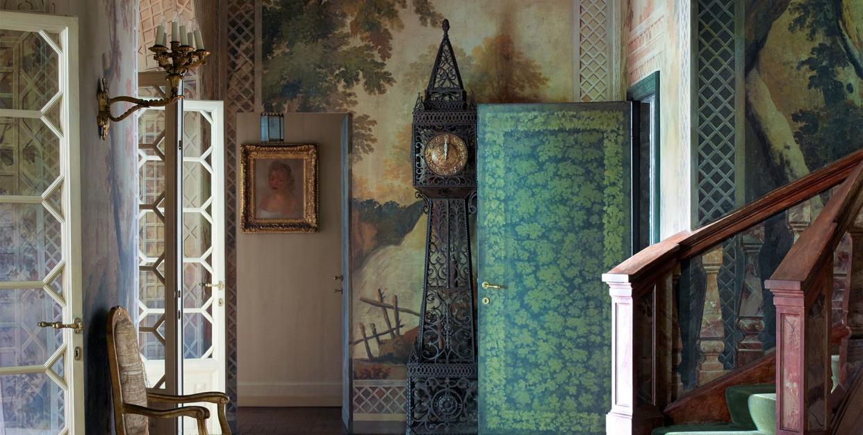 very dramatically lit entrance with a scroll like grandfather clock against the far wall and a scenic wallpaper behind it and in the foreground is a lone chair to the left and a very sweeping staircase with green runner to the right