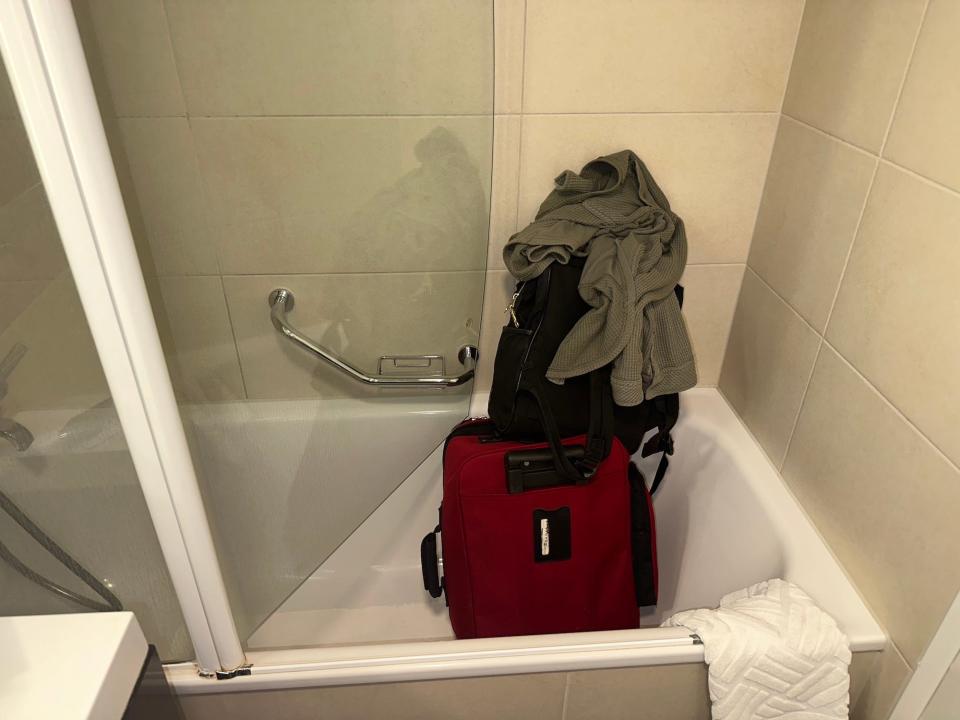 red suitcase with black backpack and green waffle shirt stacked on top inside a white bathtub