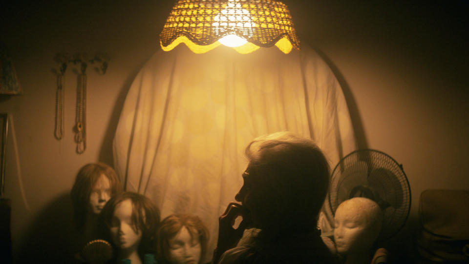 A woman stands under warm overhead light with wigs in the background in a scene from The Vigil.