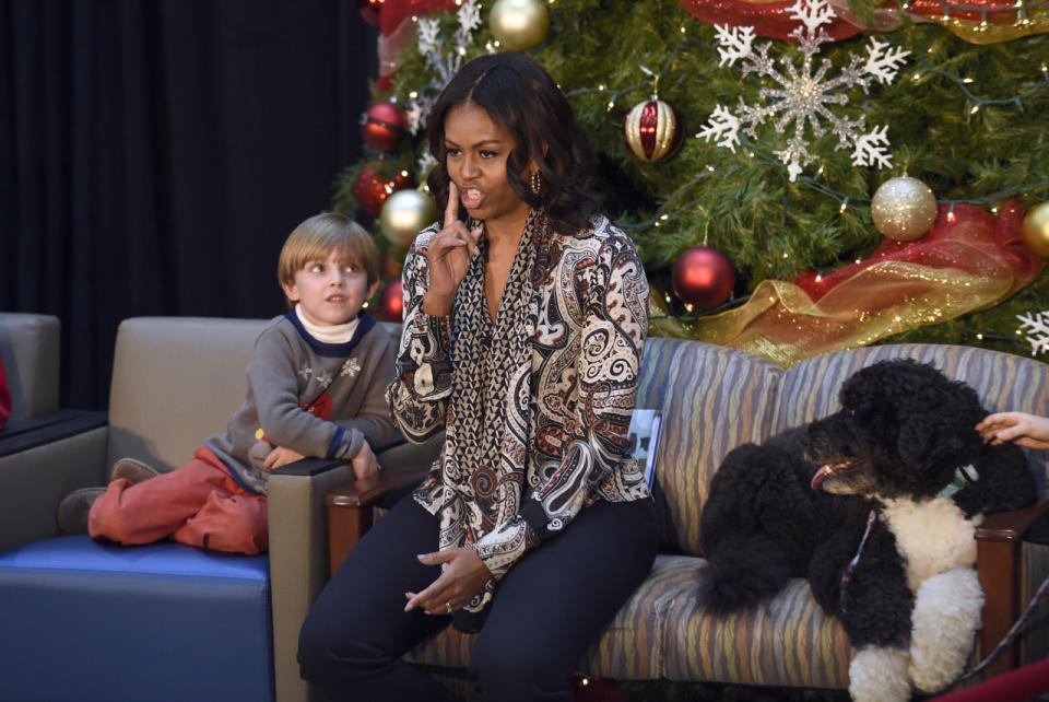 Michelle Obama in a paisley print blouse and navy blue trousers quieting the audience before reading a story to an audience of kids during a visit to the Children’s National Health System in Washington. 